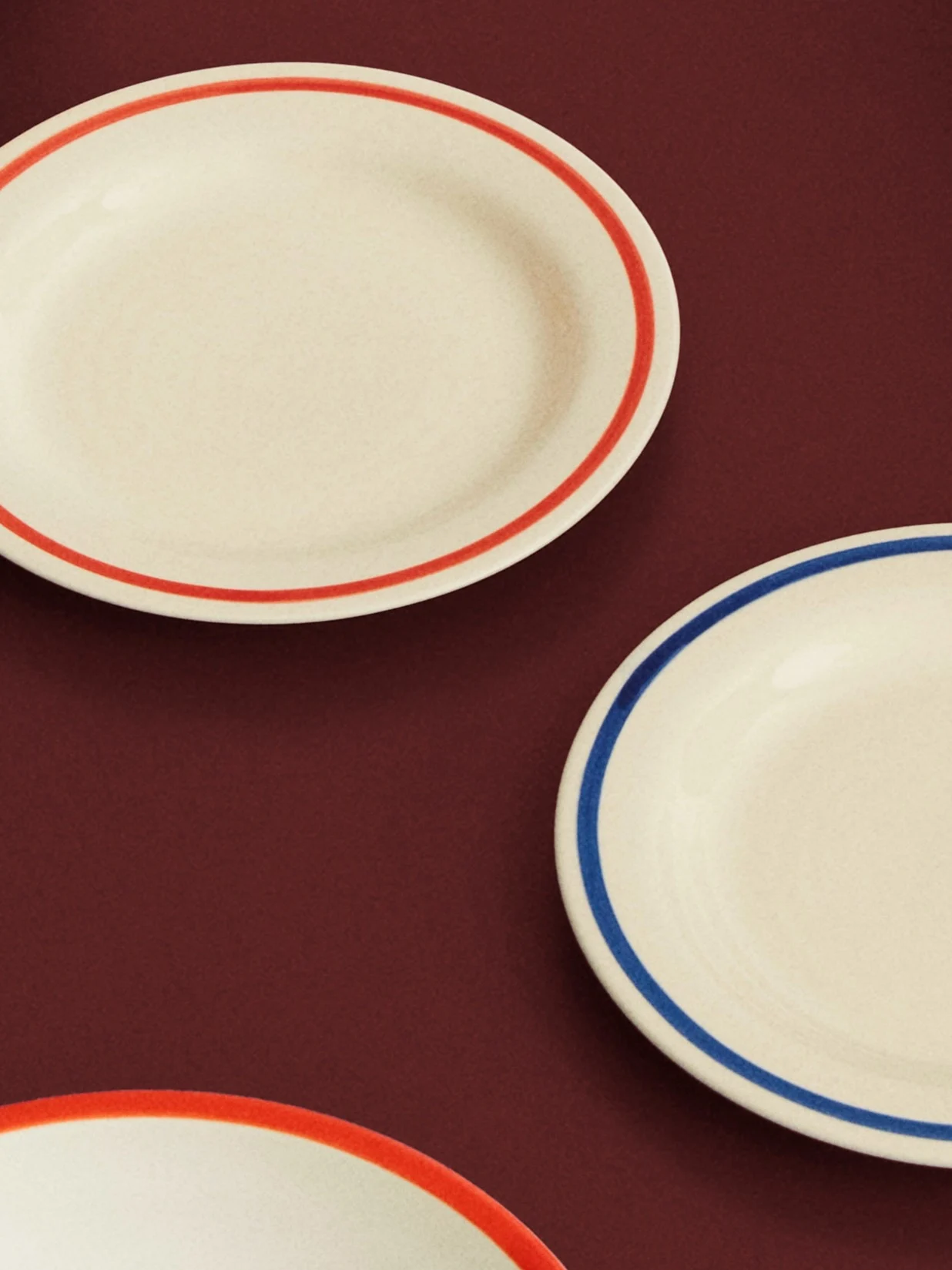 Três pratos ovais creme com bordas vermelhas e azuis sobre uma superfície vermelha escura.