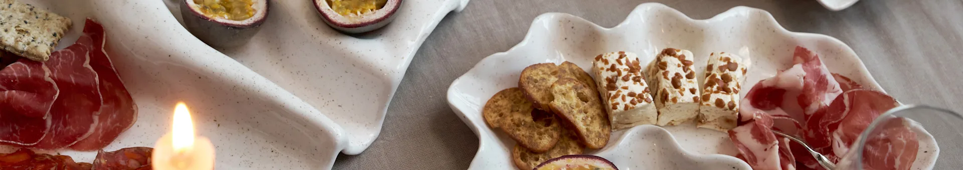 Um aperitivo em duas travessas em forma de concha com enchidos, bolachas, queijo e maracujá, com uma vela acesa.