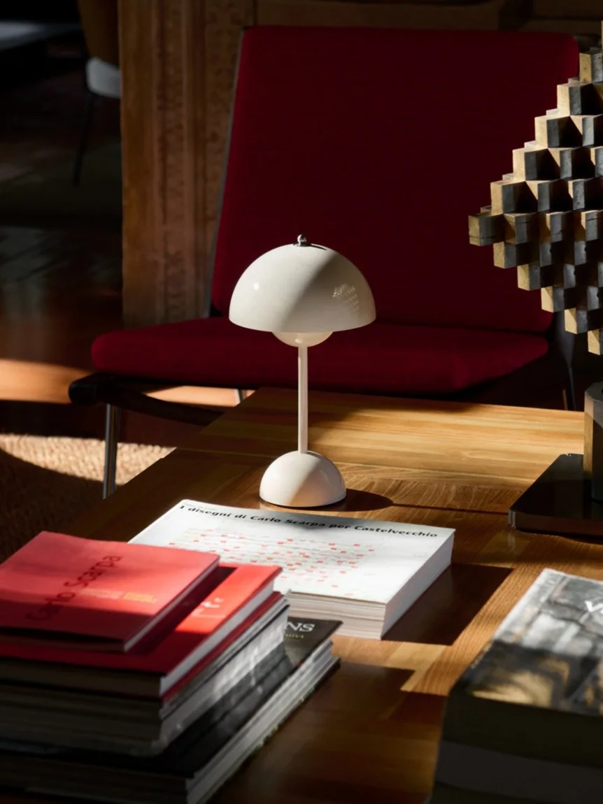 Um candeeiro de mesa branco em forma de cogumelo, livros e uma escultura geométrica numa mesa de centro de madeira, com uma poltrona vermelha atrás.