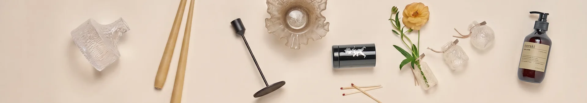 Uma coleção de artigos de decoração para casa, incluindo vasos de vidro, pauzinhos, um castiçal preto, uma tigela de vidro frisada, um recipiente preto, uma flor laranja e uma garrafa de loção para as mãos.