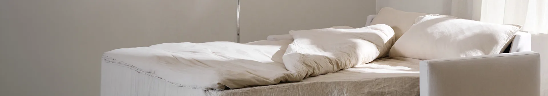 Uma cama com roupa de cama e almofadas de cor clara amassadas, suavemente iluminada por luz natural.