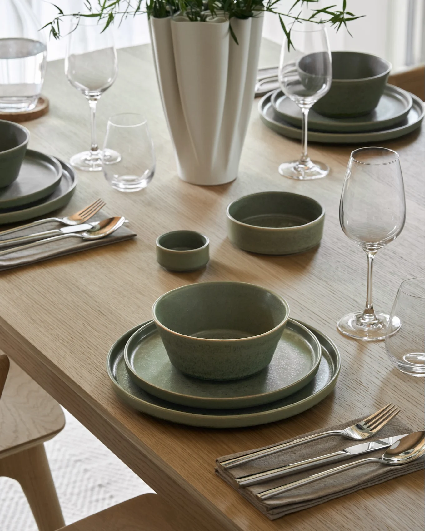 Mesa de jantar posta com tigelas de sopa cremosa, pratos a condizer, copos de vinho e água, talheres e um vaso com folhagem verde.