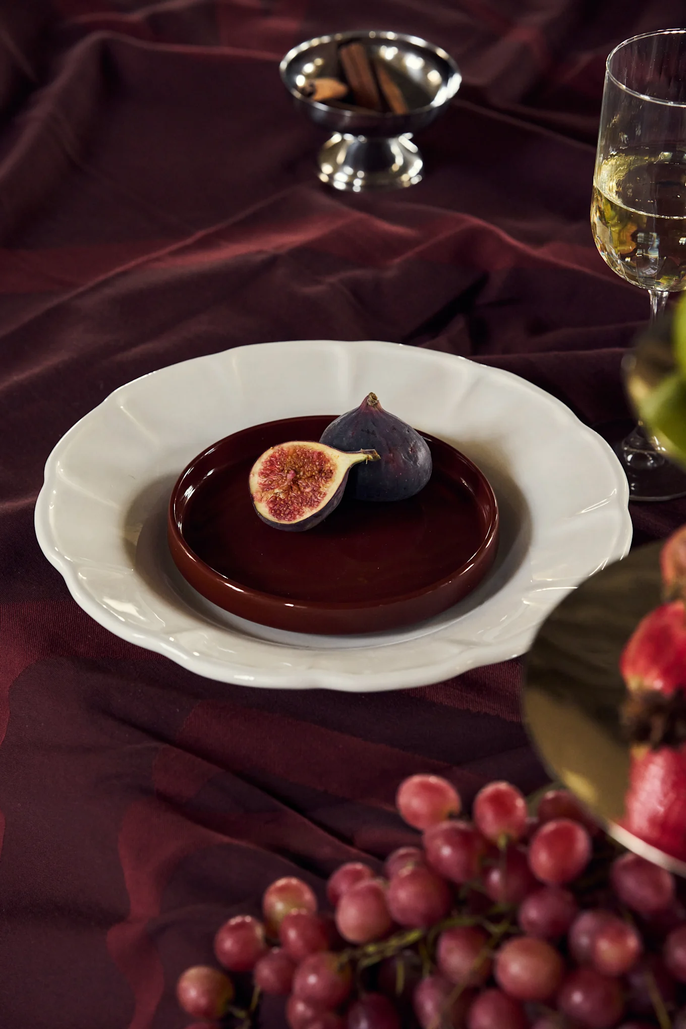 Figos num prato sobre uma toalha de mesa bordô com vinho, uvas e paus de canela.