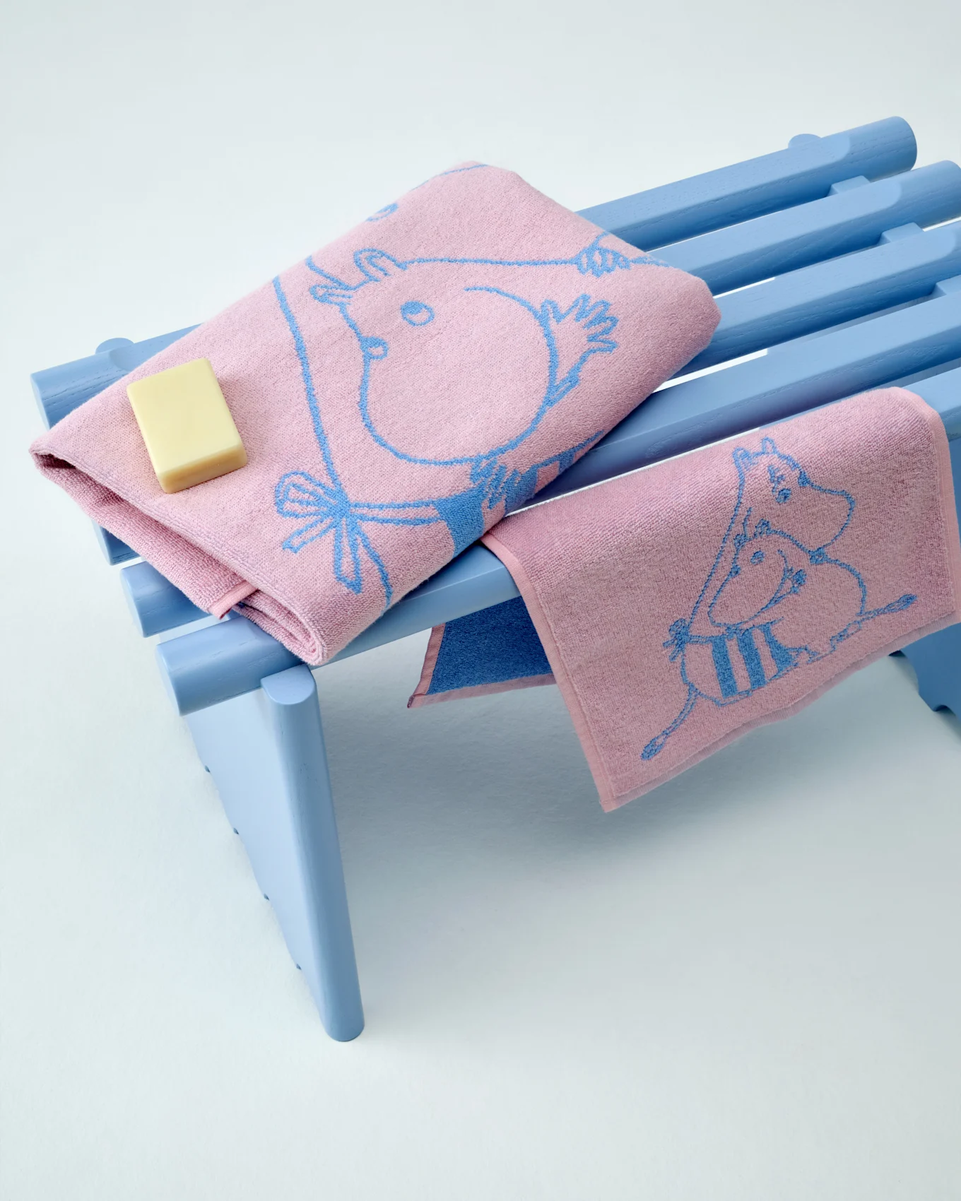 Duas toalhas Moomin cor-de-rosa com padrões azuis, uma dobrada com uma barra de sabão, sobre um banco azul claro.