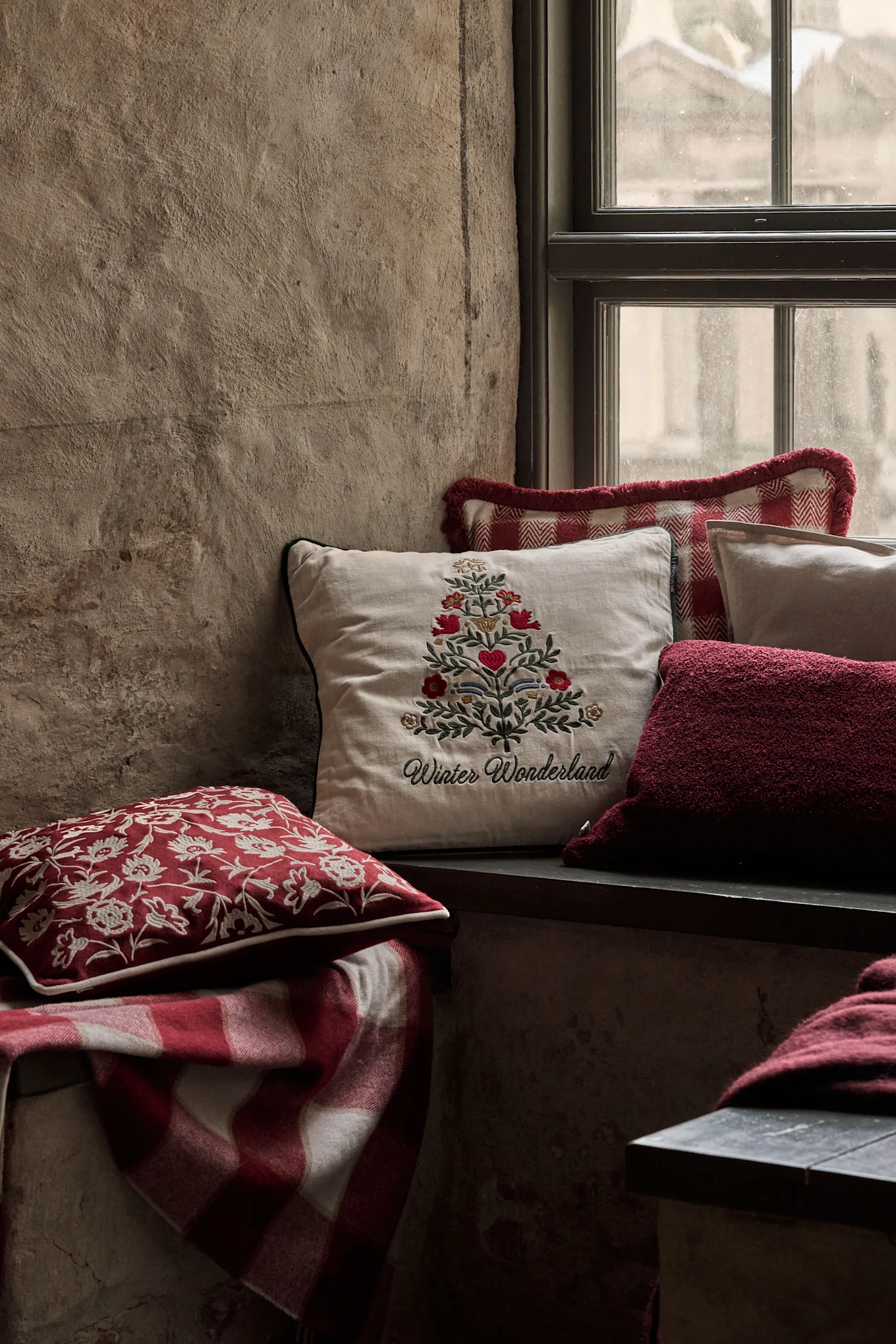 Aconchegante recanto de janela com almofadas festivas, incluindo uma com árvore de Natal bordada e 'Winter Wonderland', e uma manta xadrez vermelha.