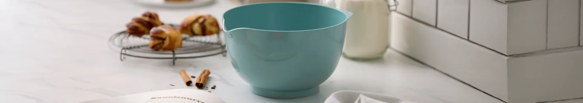 Tigela azul clara, doces na grelha, garrafa de leite e livro de receitas em bancada de mármore.