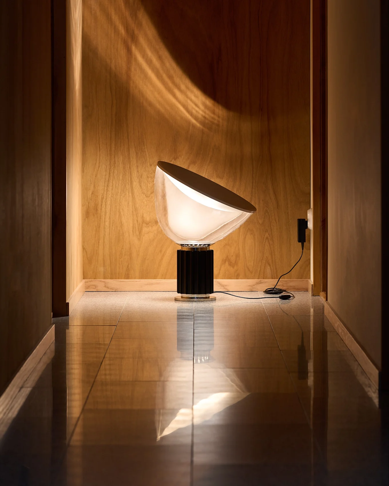 Candeeiro moderno com abajur branco opaco semicircular e base preta canelada em corredor de madeira com chão refletor. Padrão de luz na parede.