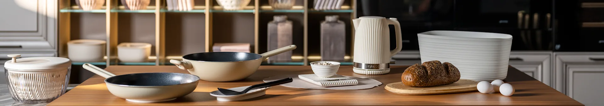 Bancada de cozinha com frigideiras bege, uma chaleira branca canelada, um pão de sementes numa tábua e ovos. Acessórios de cozinha modernos.