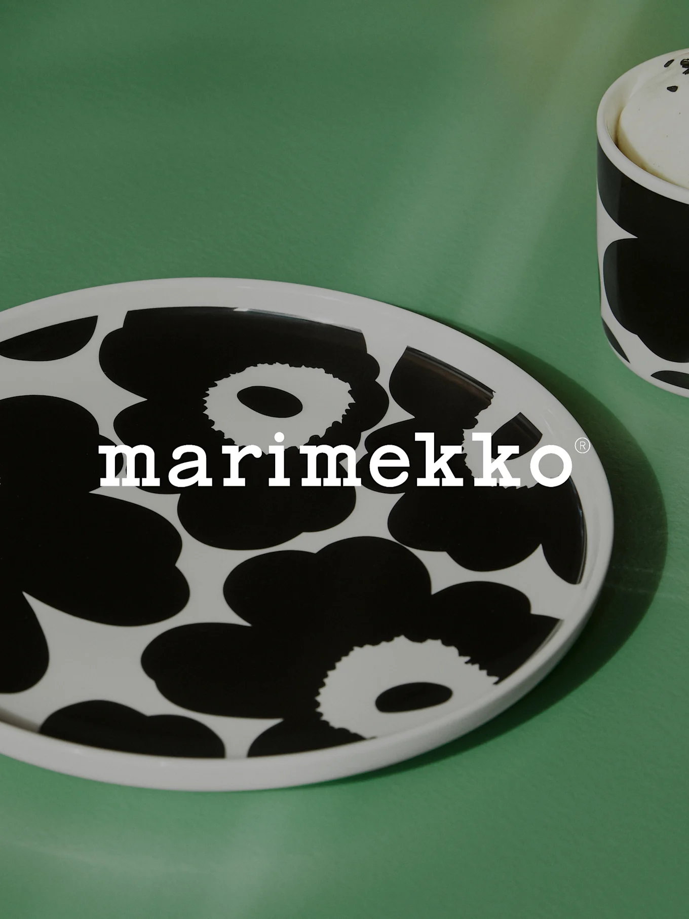 Prato oval branco com padrão floral preto e logótipo Marimekko numa superfície verde, com uma caneca correspondente parcialmente visível.