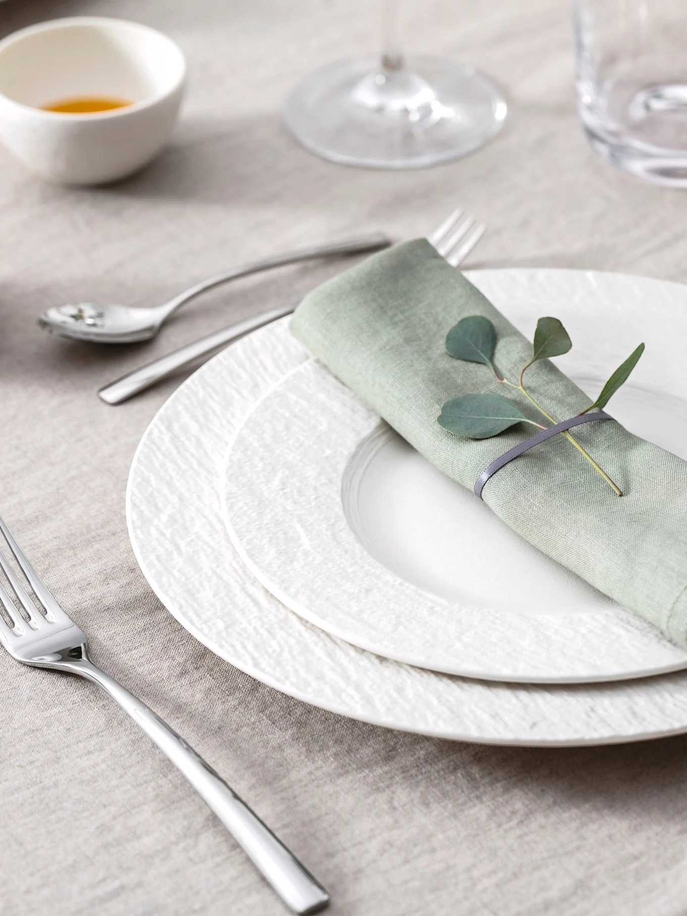 Elegante mesa posta com guardanapo de linho verde claro, folhas de eucalipto, pratos brancos texturizados e talheres sobre uma toalha de mesa bege.