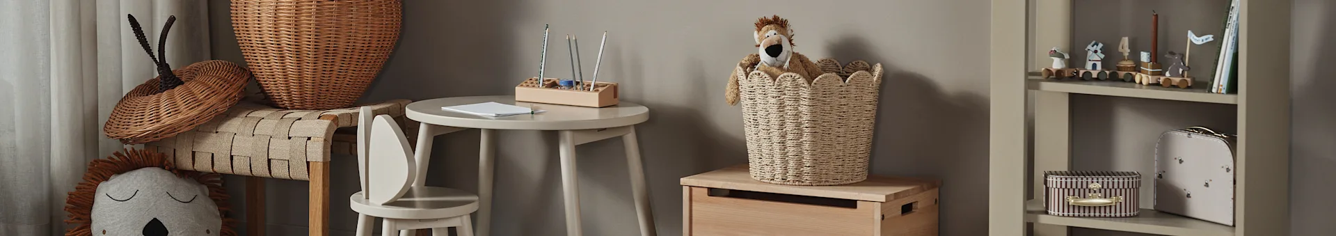 Um quarto de criança com um brinquedo de peluche de leão num cesto de verga, uma pequena mesa de madeira com lápis e uma estante com brinquedos e livros.