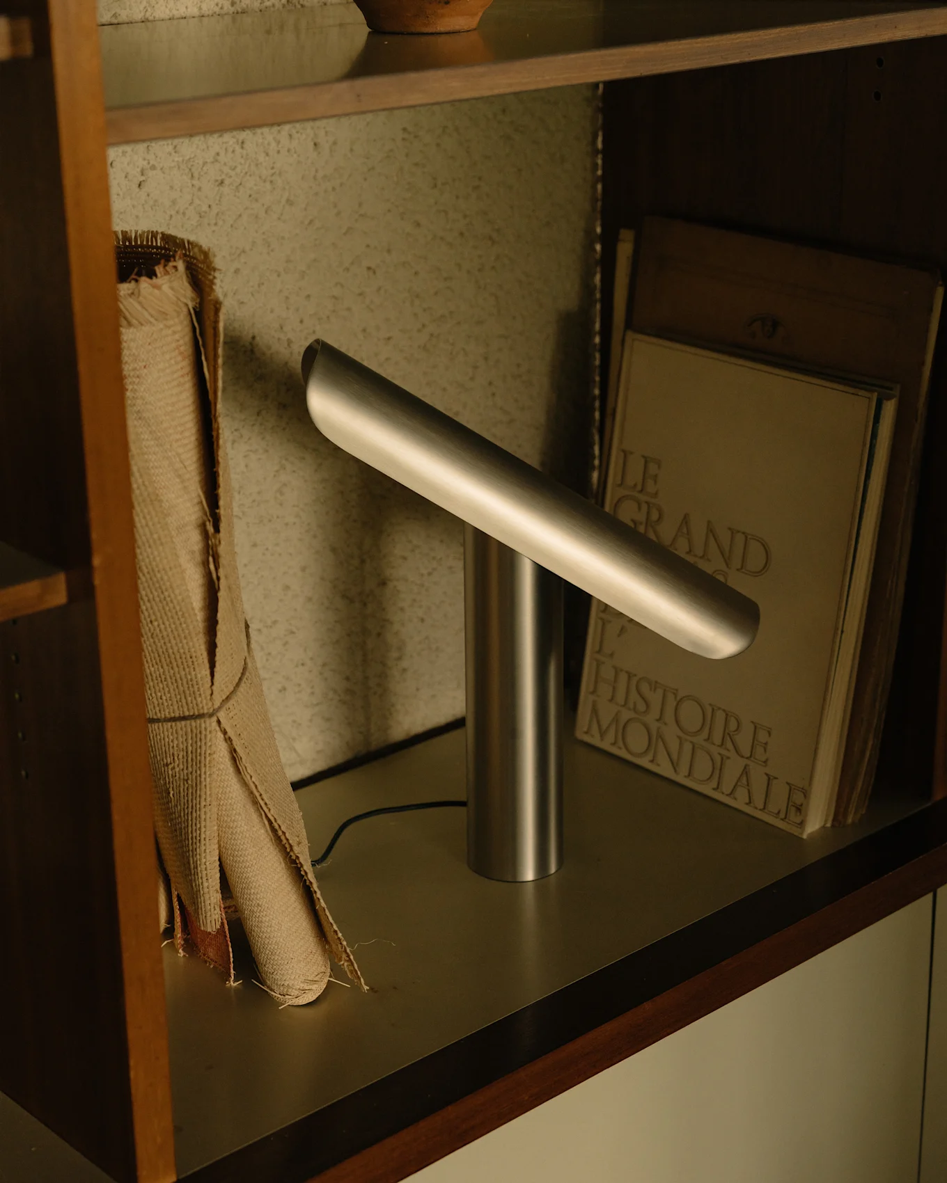 Candeeiro de secretária moderno prateado com base cilíndrica e barra de luz horizontal angulada numa prateleira.