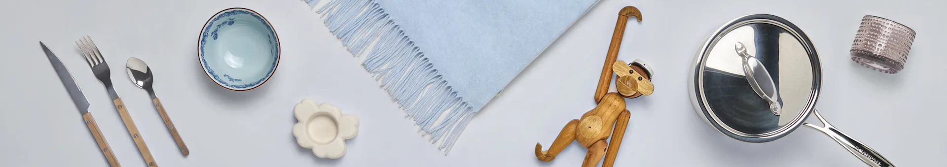 Sortimento de artigos para o lar: talheres com cabos de madeira, tigela azul, descanso de copo de flor, manta azul, macaco de madeira, panela e copo.