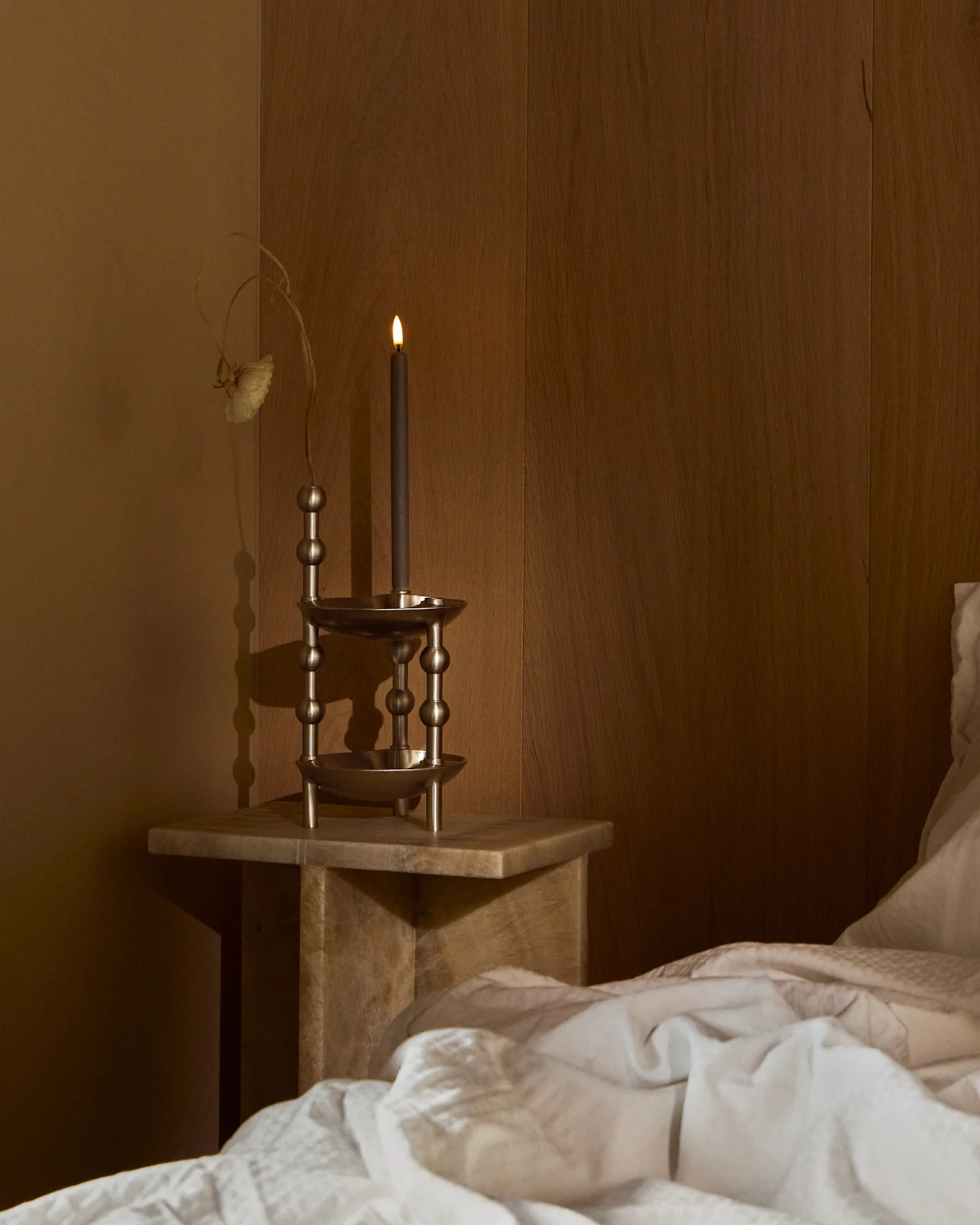 Uma vela escura acesa num castiçal de metal ornamentado sobre uma mesa de cabeceira de madeira, ao lado de uma cama com roupa de cama branca num quarto escuro.