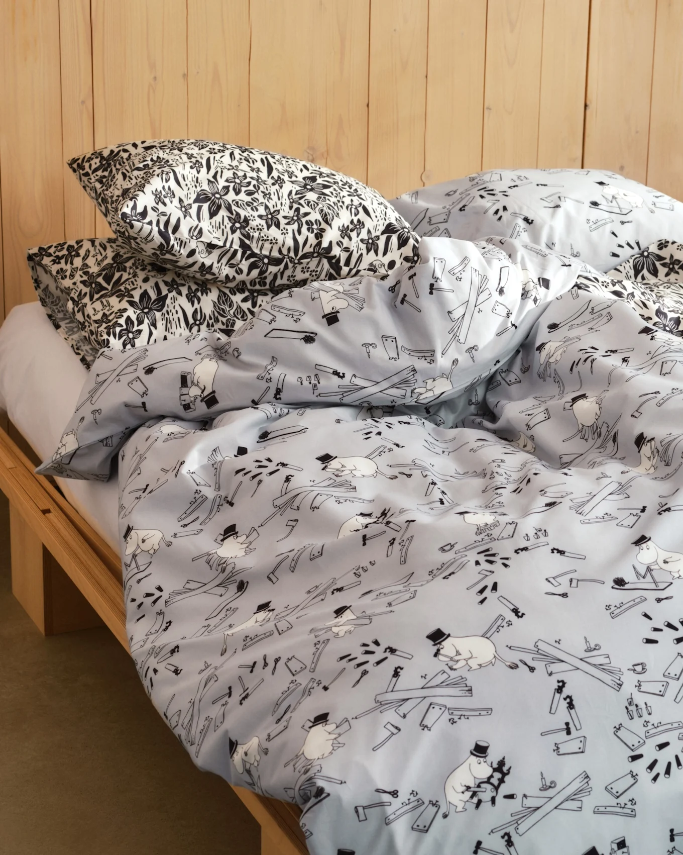 Cama com almofadas de estampa floral preta e branca e edredom azul claro com figuras de desenhos animados, ferramentas e pranchas de madeira. Estrutura de cama de madeira.