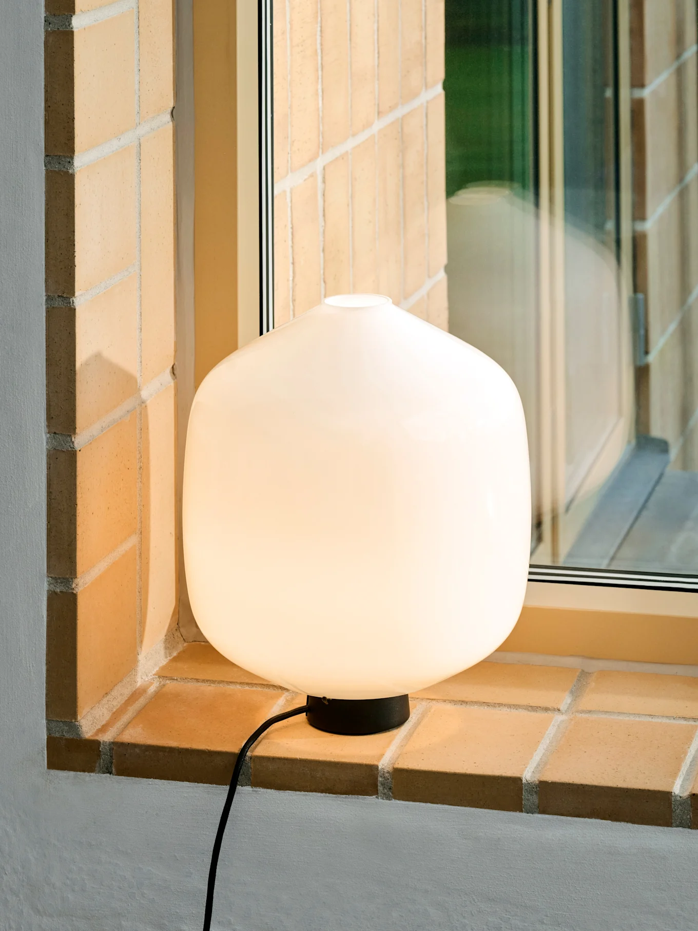 Uma luminosa e arredondada lâmpada de mesa branca com base e cabo pretos sobre um parapeito de tijolo.