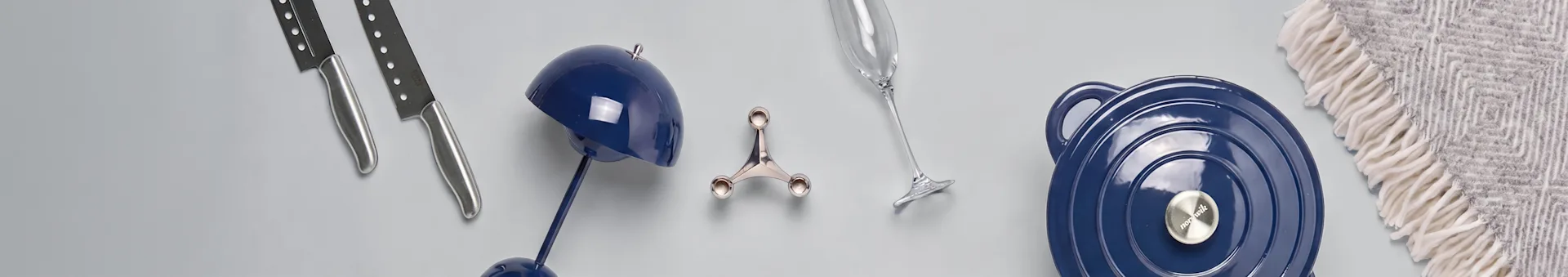 Duas facas, um candeeiro azul, um suporte prateado, uma flûte de champanhe, uma panela azul e uma manta cinzenta estampada numa superfície clara.