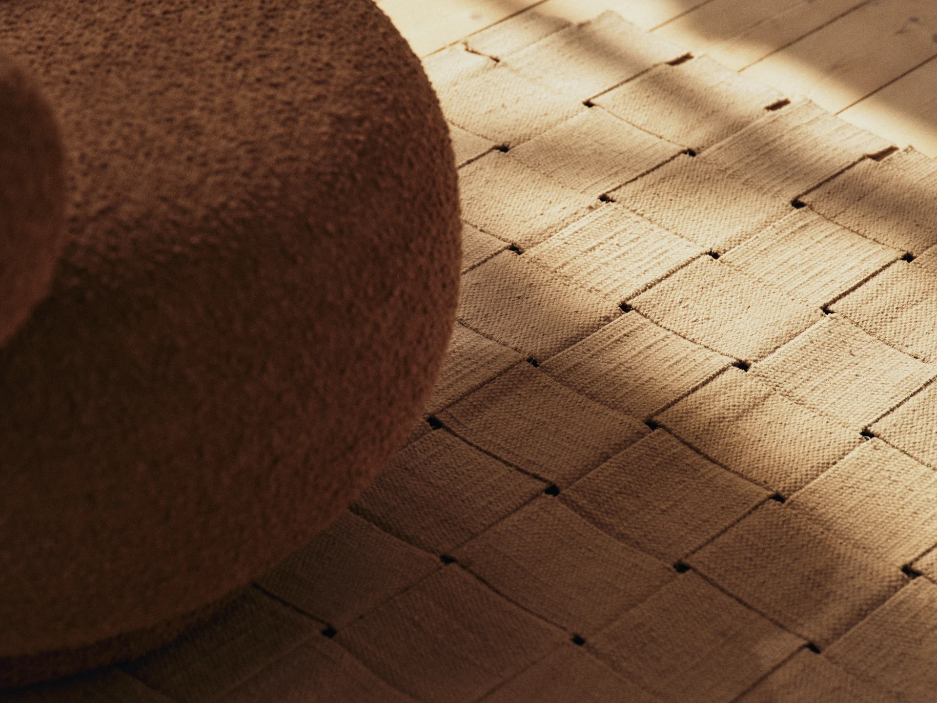 Móveis bouclé marrons, um tapete tecido e um piso de madeira com sombras ensolaradas.