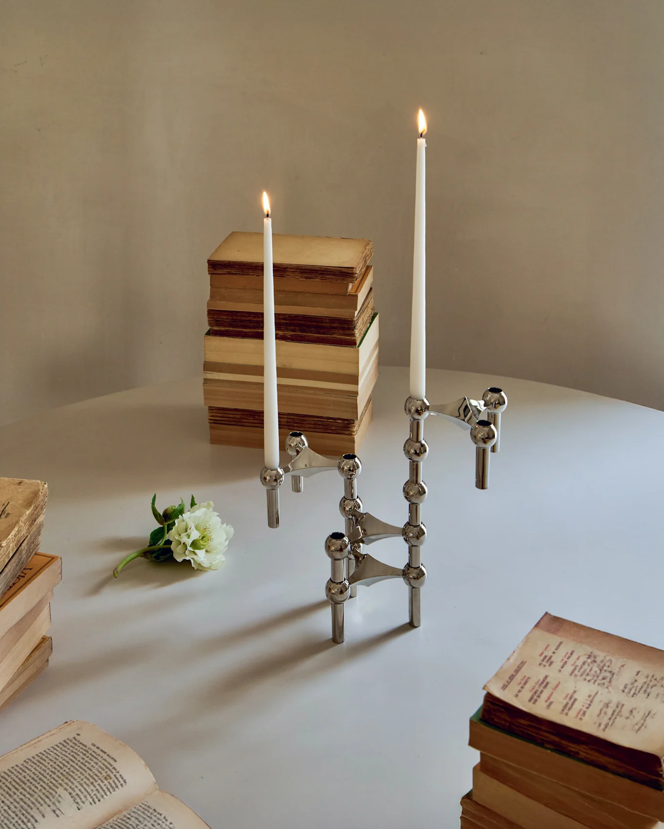 Dois castiçais modulares cromados com velas brancas acesas, rodeados por livros antigos e uma flor branca numa mesa.