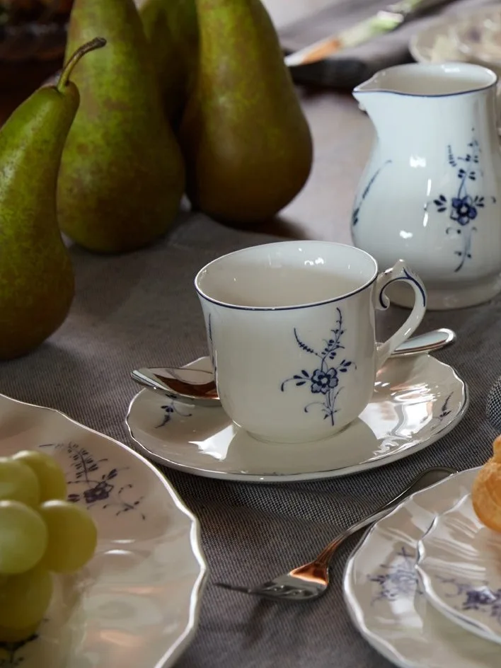 Xícara, pires e jarra de porcelana branca com padrão floral azul em mesa com peras e uvas.