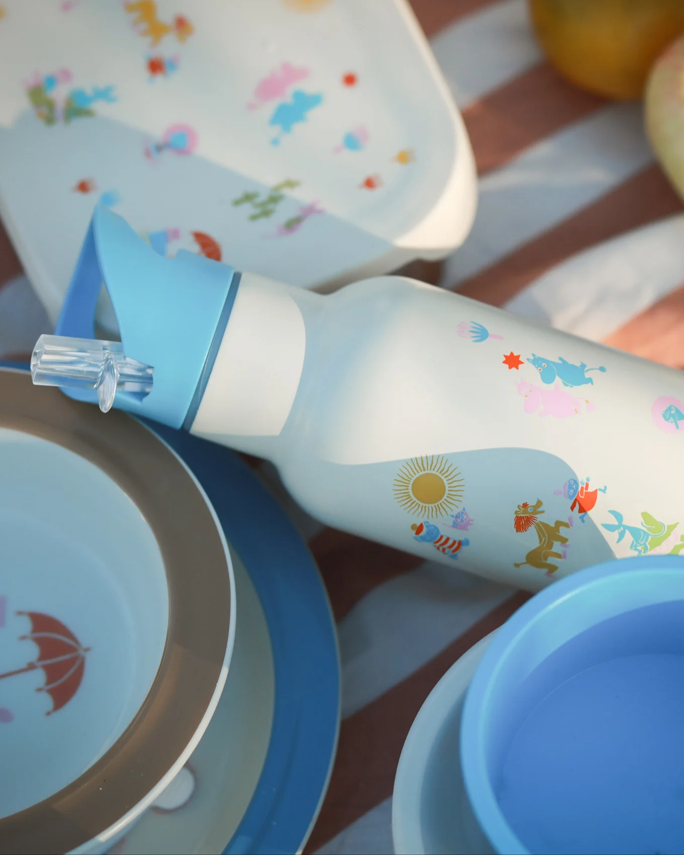 Garrafa de água branca com palhinha azul, personagens de desenhos animados coloridos e sol, rodeada por pratos de piquenique a condizer sobre um cobertor às riscas.