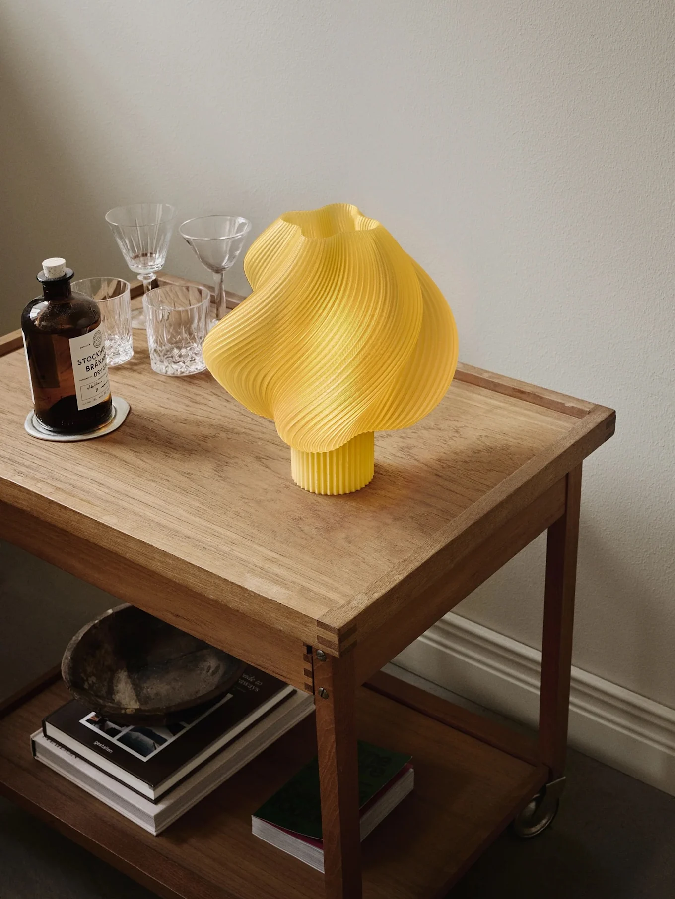 Candeeiro plissado amarelo num carrinho de madeira com garrafa de gin, copos e livros na prateleira inferior.