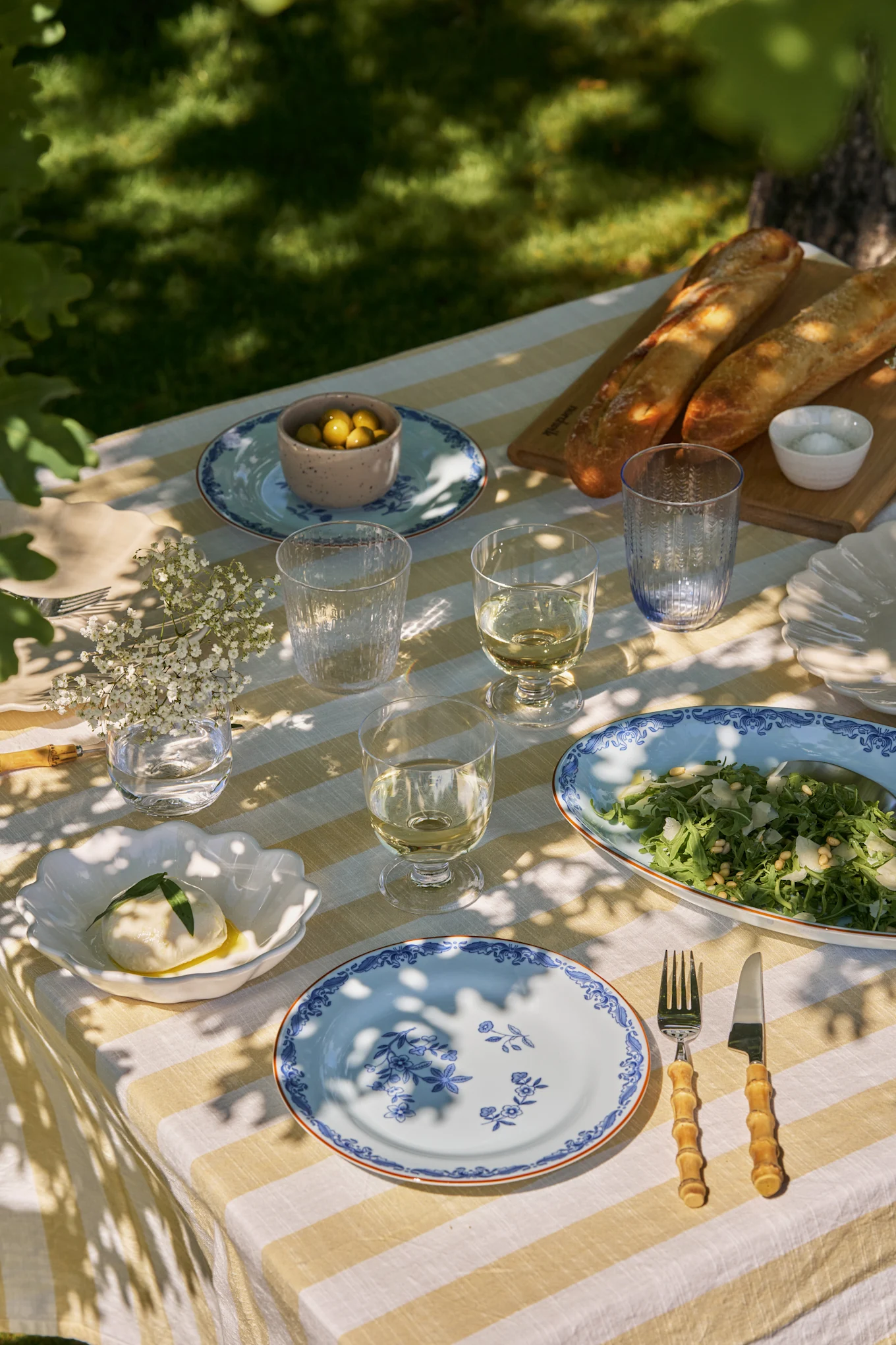 Mesa de jantar ao ar livre com toalha de mesa listrada branca e amarela, pratos florais azuis, vinho, salada, mozzarella, azeitonas e baguetes.