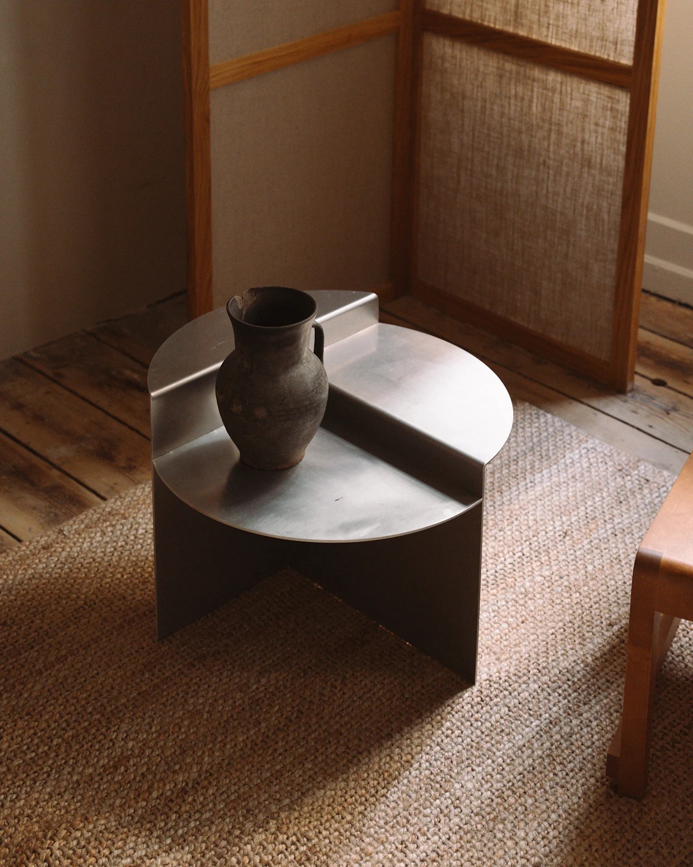 Uma mesa de centro minimalista, baixa e redonda, de metal prateado, com um vaso rústico de cerâmica escura sobre um tapete tecido.