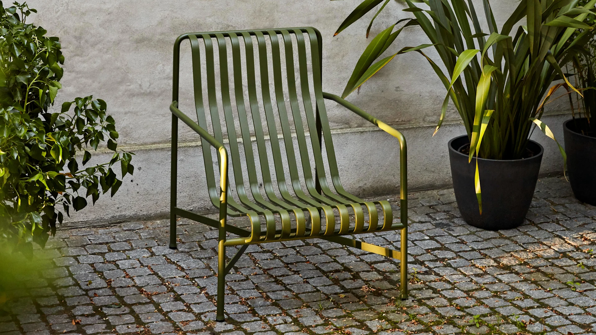 Cadeira lounge de exterior de metal com ripas verde-escura num pátio de paralelepípedos, emoldurada por plantas verdes.