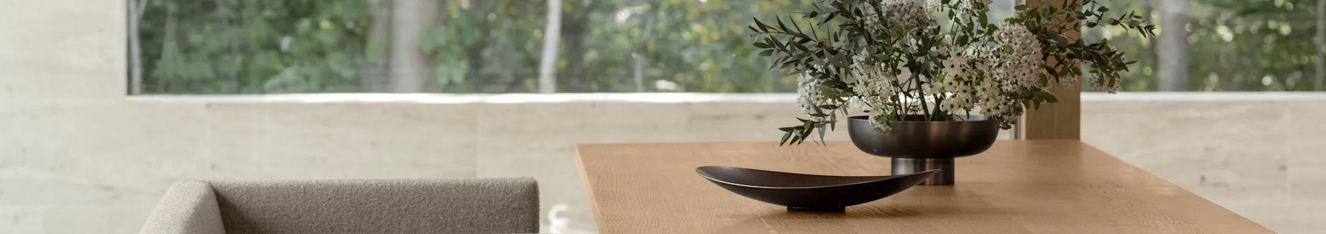 Mesa de jantar moderna de madeira com uma tigela decorativa escura e um vaso de flores brancas e verdes, com uma vista exterior verde embaçada através de uma grande janela.