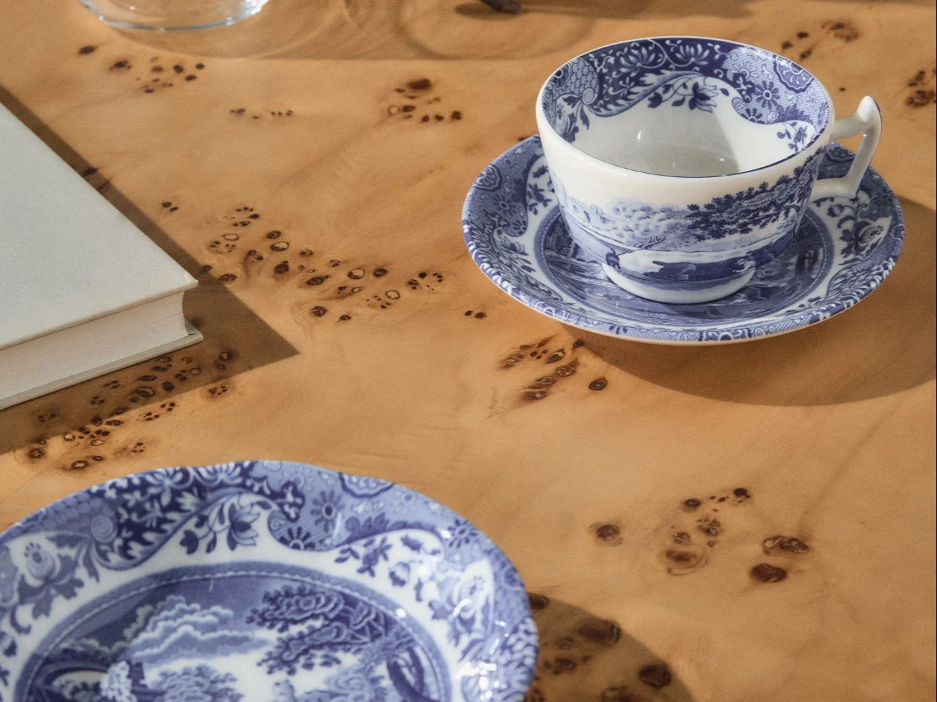 Xícara de chá e pires com padrão azul e branco com um prato a condizer sobre uma mesa de madeira clara com nós, um livro branco visível ao fundo.