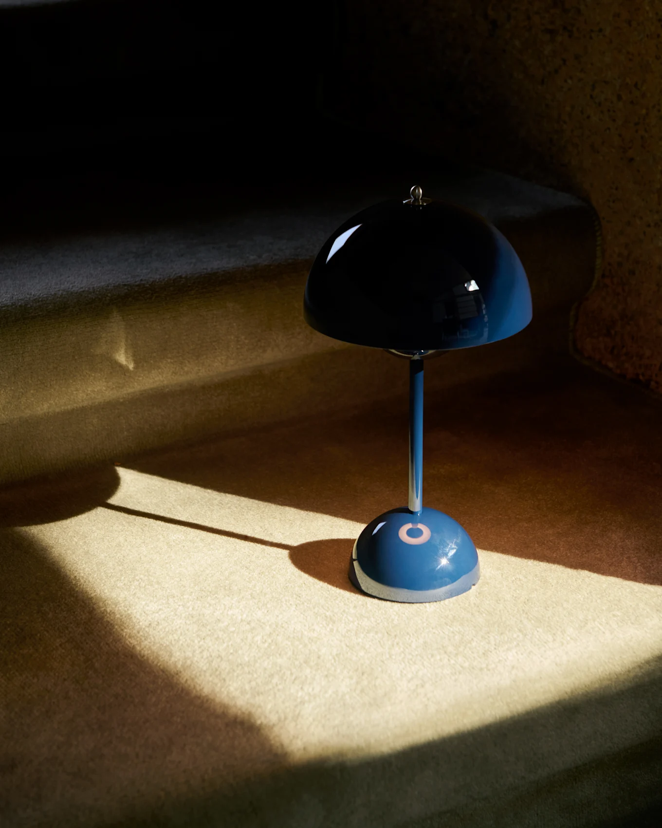 Um candeeiro de mesa azul em forma de cogumelo com um abajur refletor está sobre uma superfície alcatifada, parcialmente iluminado pelo sol.