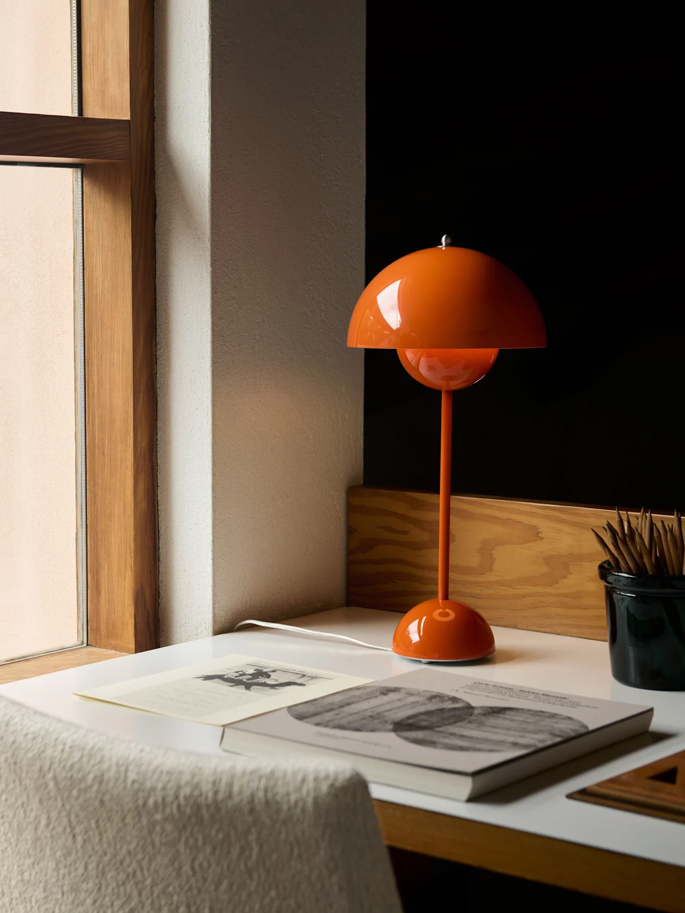 Um candeeiro de mesa laranja em forma de cogumelo numa secretária branca junto a uma janela, com livros e lápis.