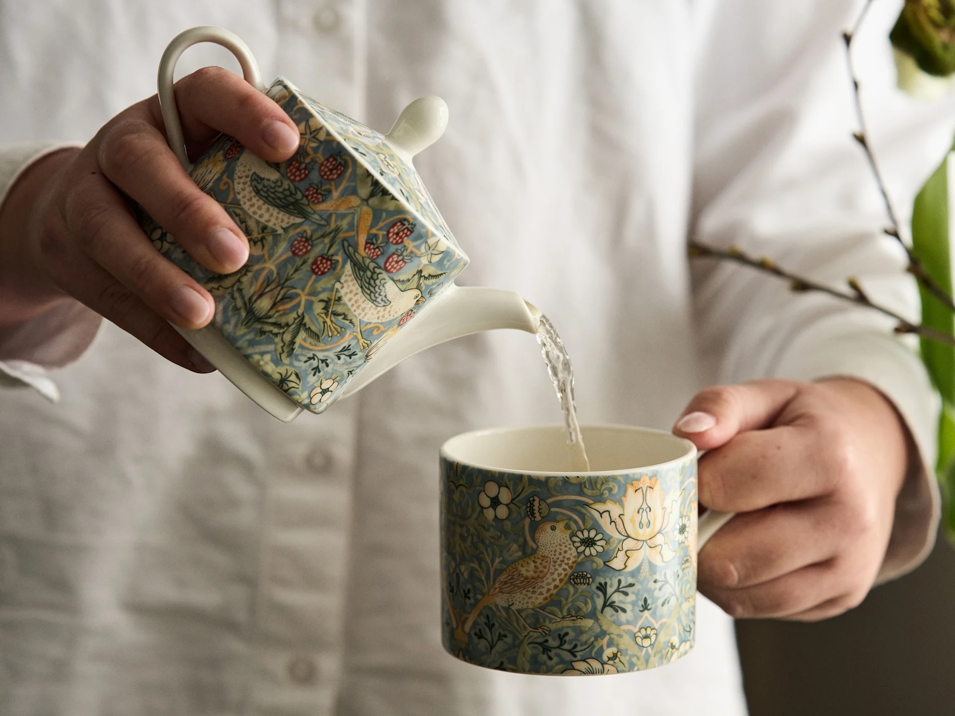 Mãos em camisa branca a servir de um bule quadrado estampado para uma caneca a condizer com design de pássaros e flores.