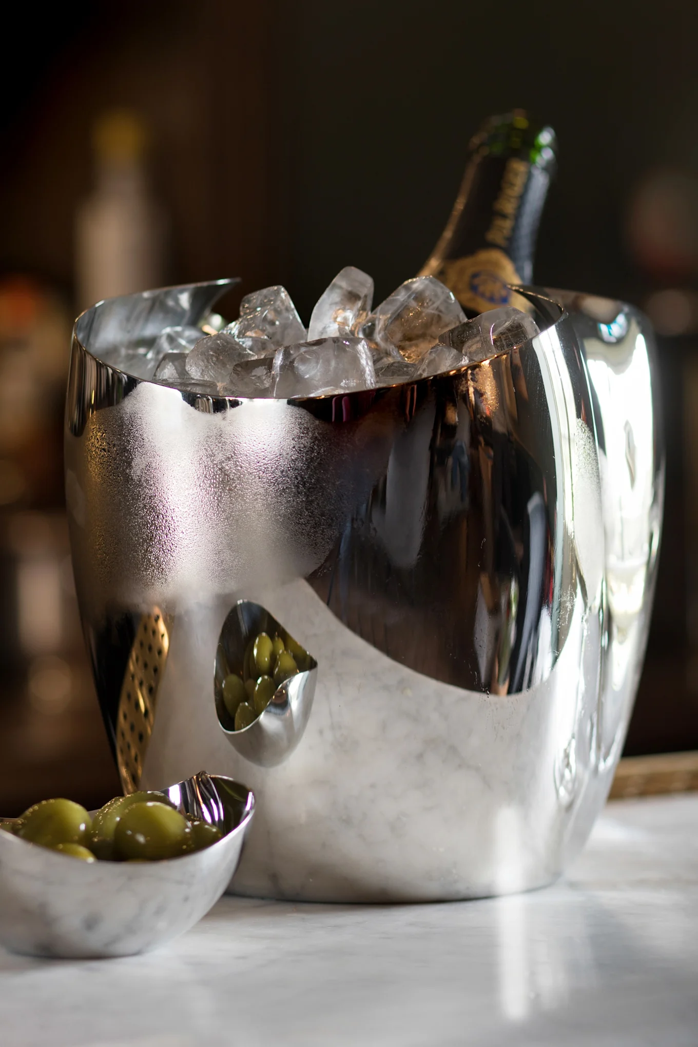 Um balde de gelo prateado reflexivo com uma garrafa de champanhe e gelo, ao lado de duas pequenas tigelas de azeitonas verdes sobre mármore.