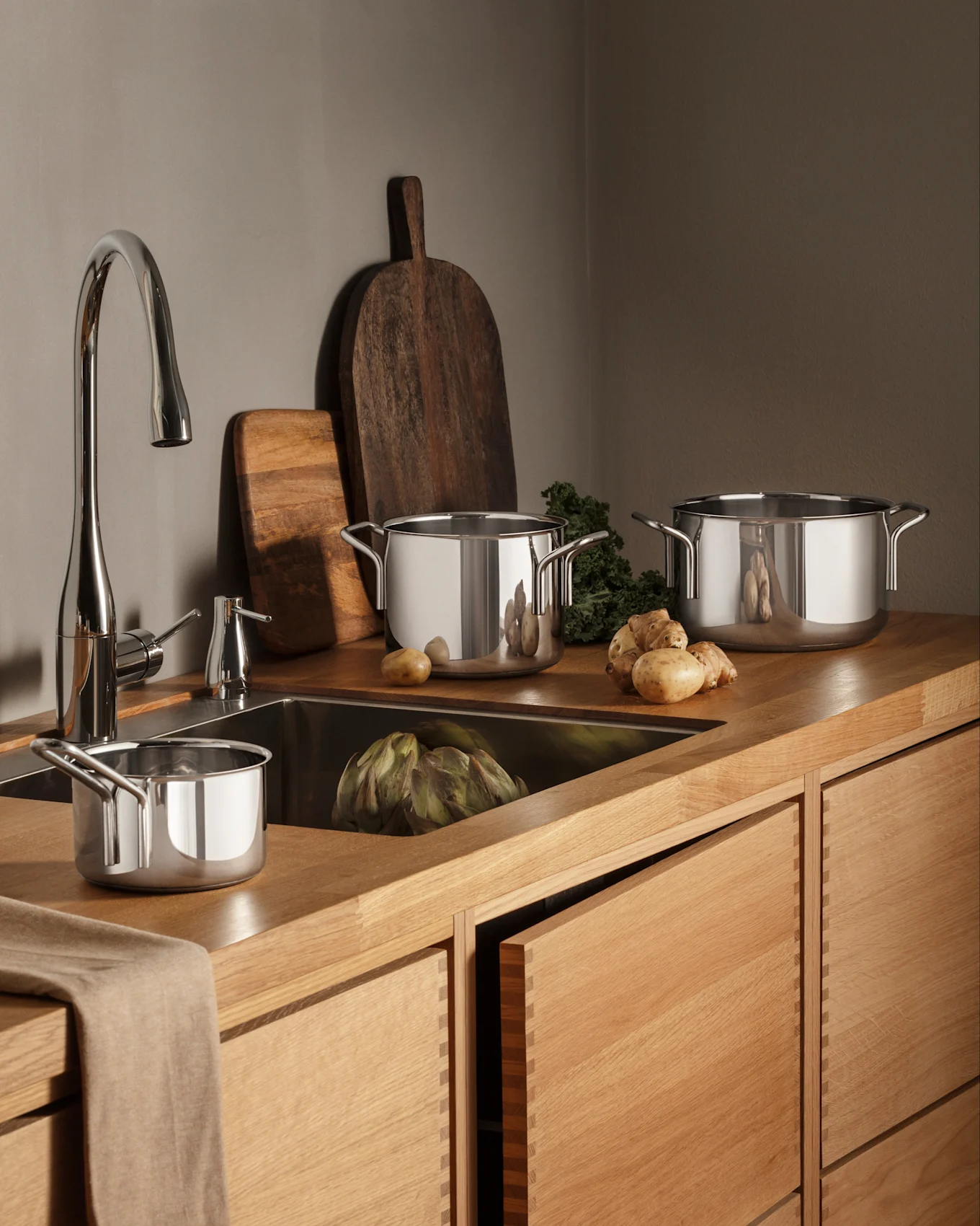 Uma cozinha moderna com bancadas e armários de madeira, uma torneira cromada e panelas de aço inoxidável, produtos frescos, incluindo alcachofra e batatas.