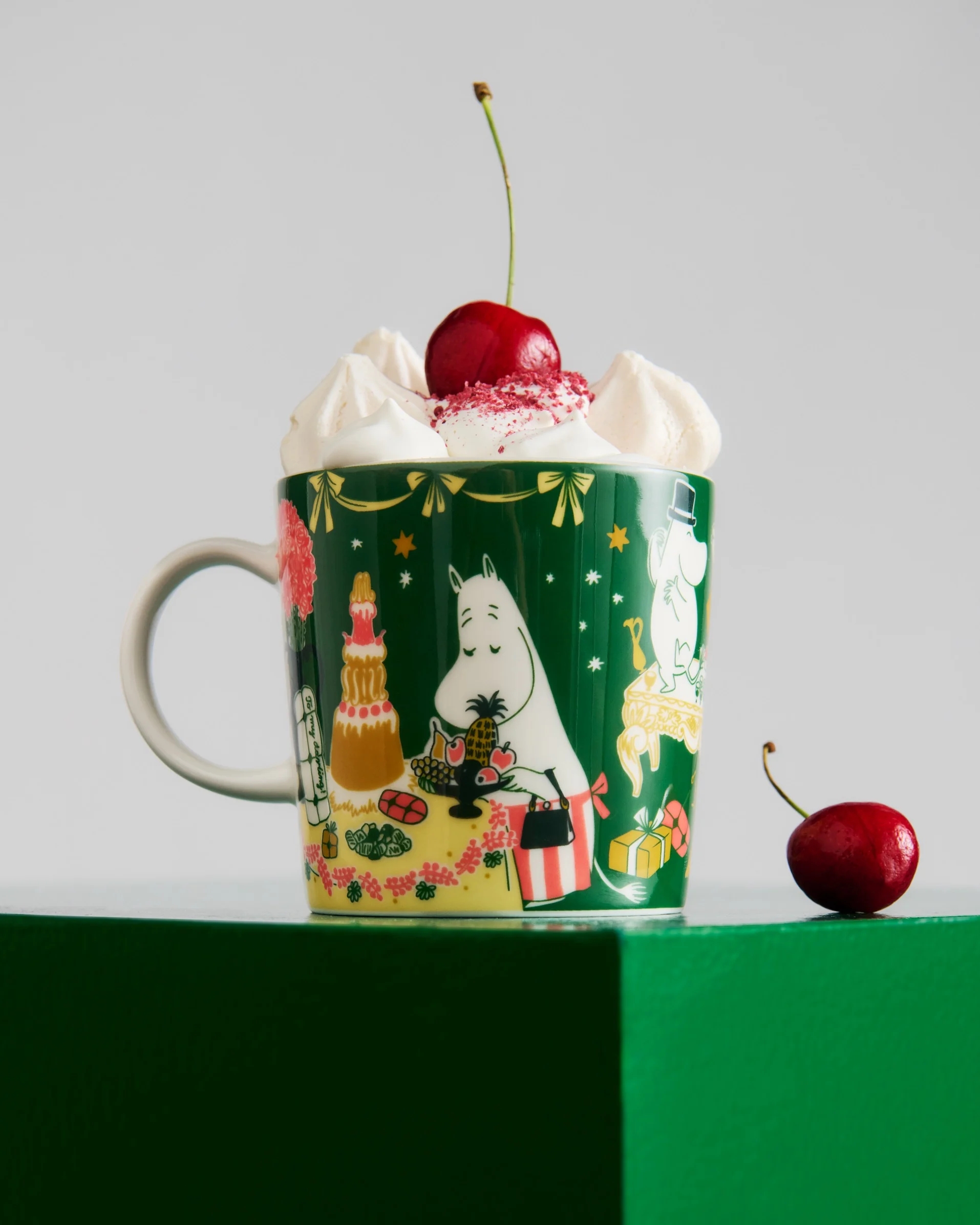 Caneca verde escura com personagens Moomin, coberta com chantilly, granulado vermelho e uma cereja. Outra cereja está na superfície verde.