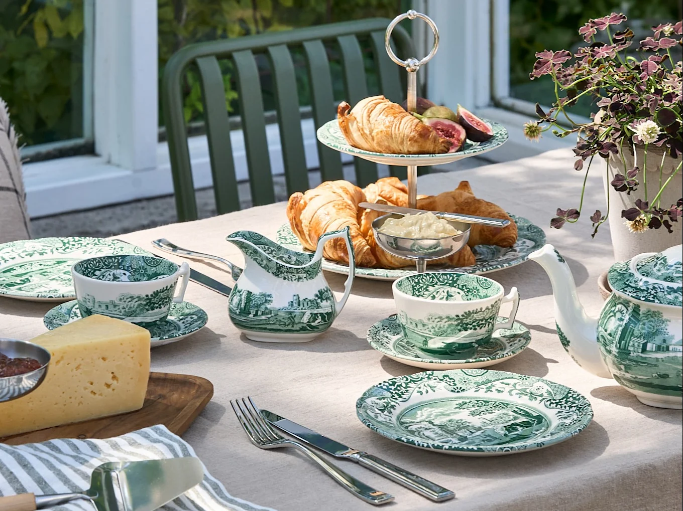 Uma mesa ao ar livre posta com louça de padrão verde e branco, croissants, queijo e um bule, num cenário de jardim.