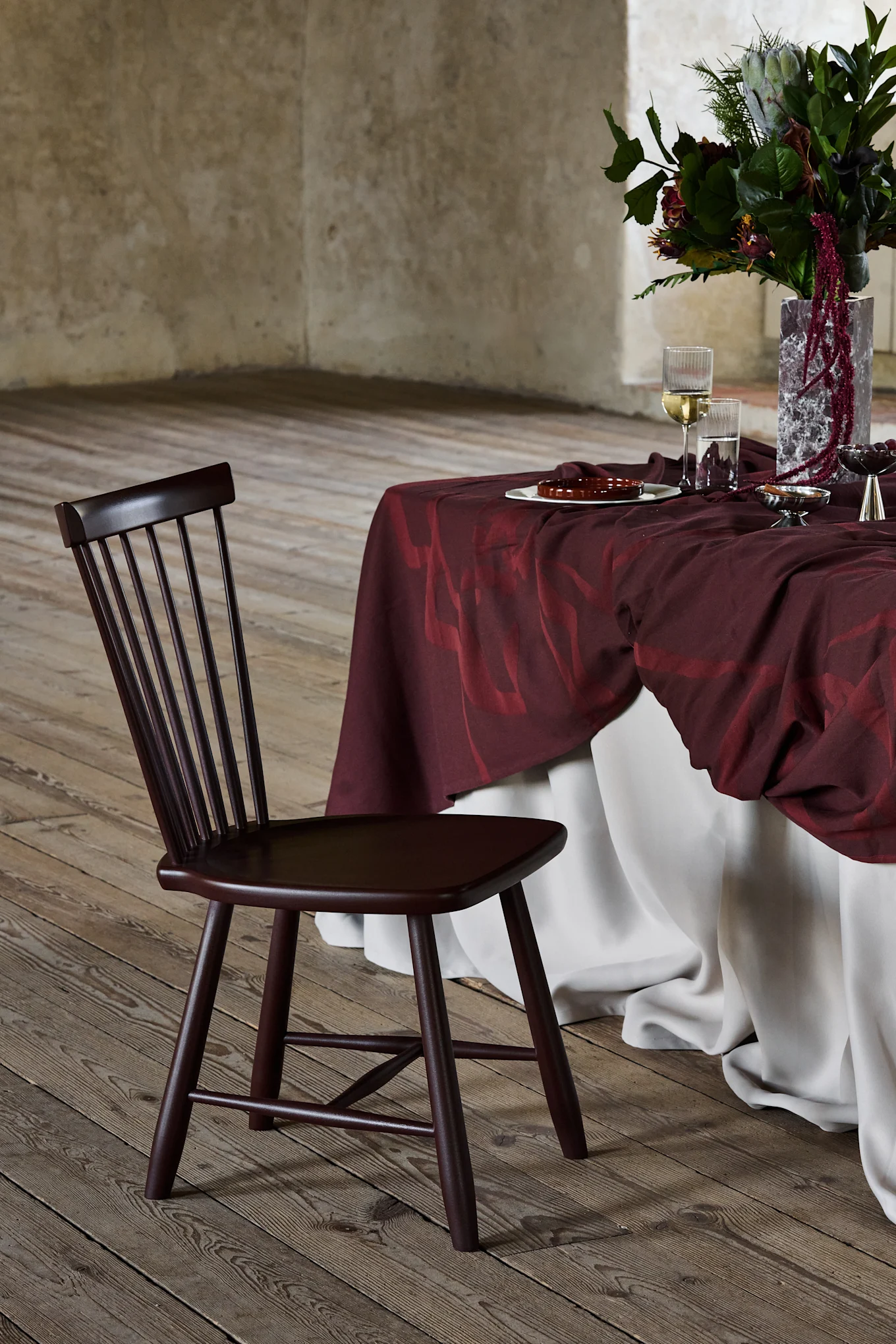 Cadeira de madeira marrom escura com encosto de ripas, ao lado de uma mesa coberta de bordô com arranjo floral, em piso de madeira.