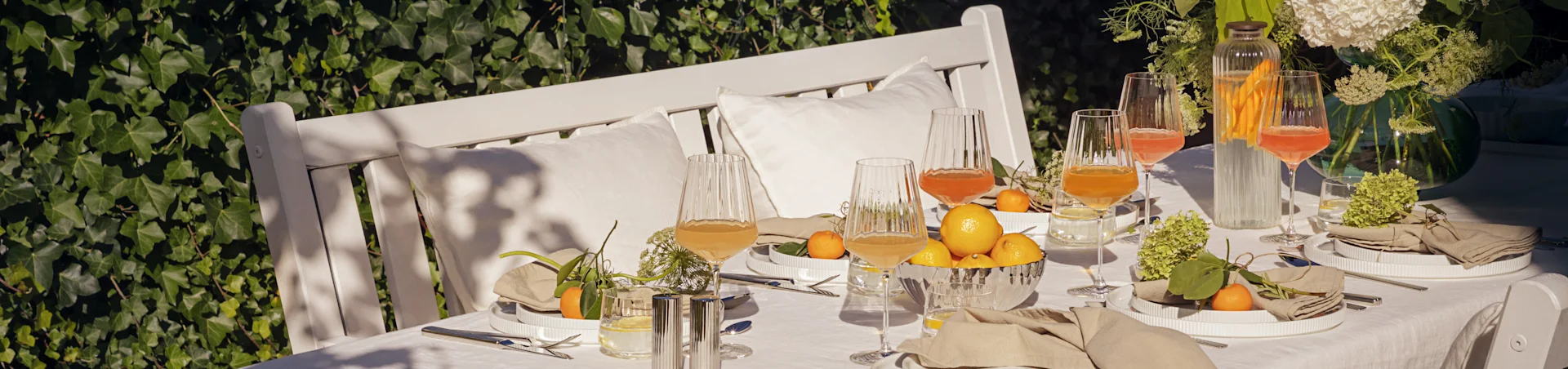 Mesa exterior posta para uma refeição de verão com pratos brancos estriados, guardanapos de linho, copos estriados, limões e mandarinas.