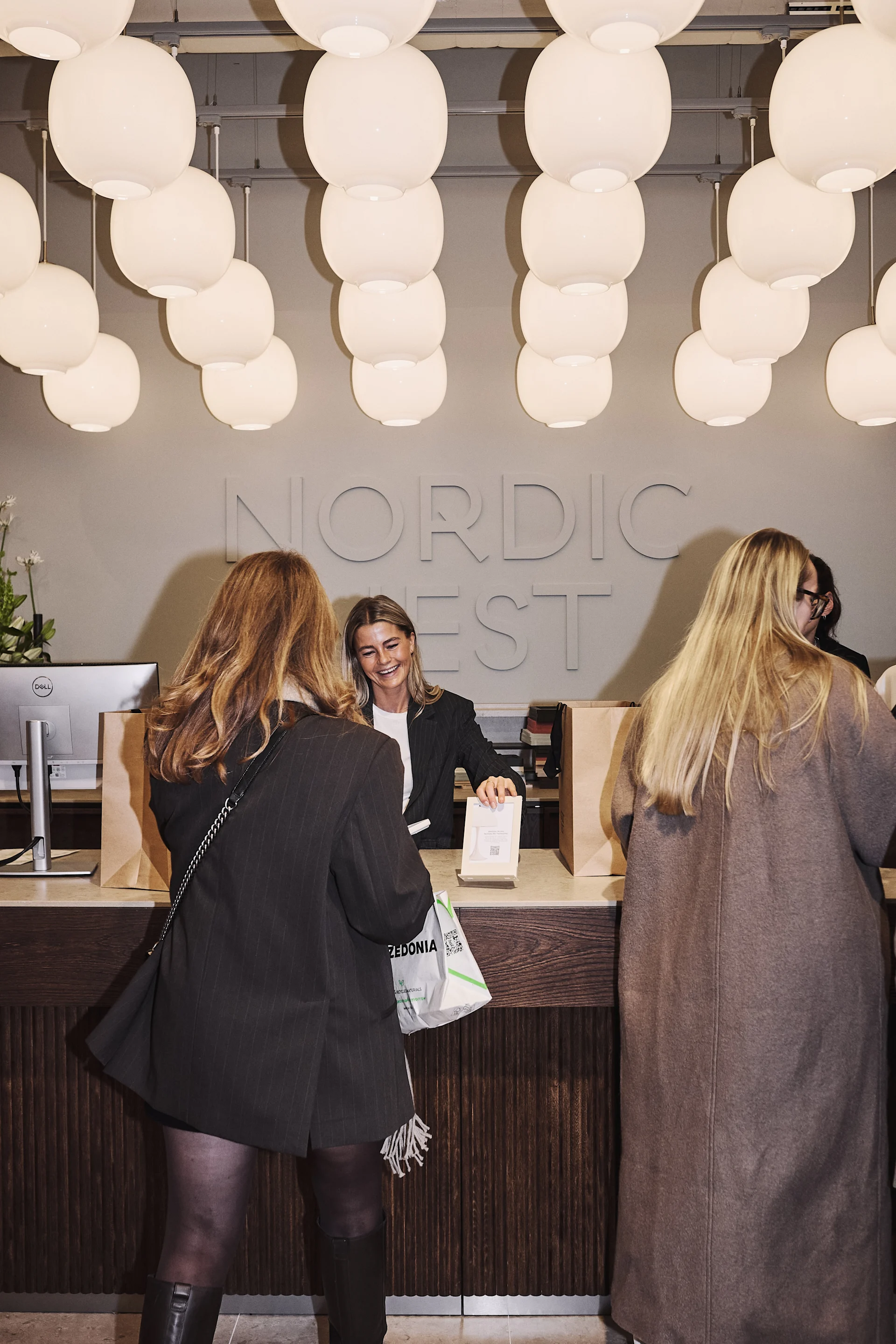 Vendedora sorri para dois clientes em balcão com sacolas, sob luminárias esféricas brancas e letreiro "NORDIC EST".