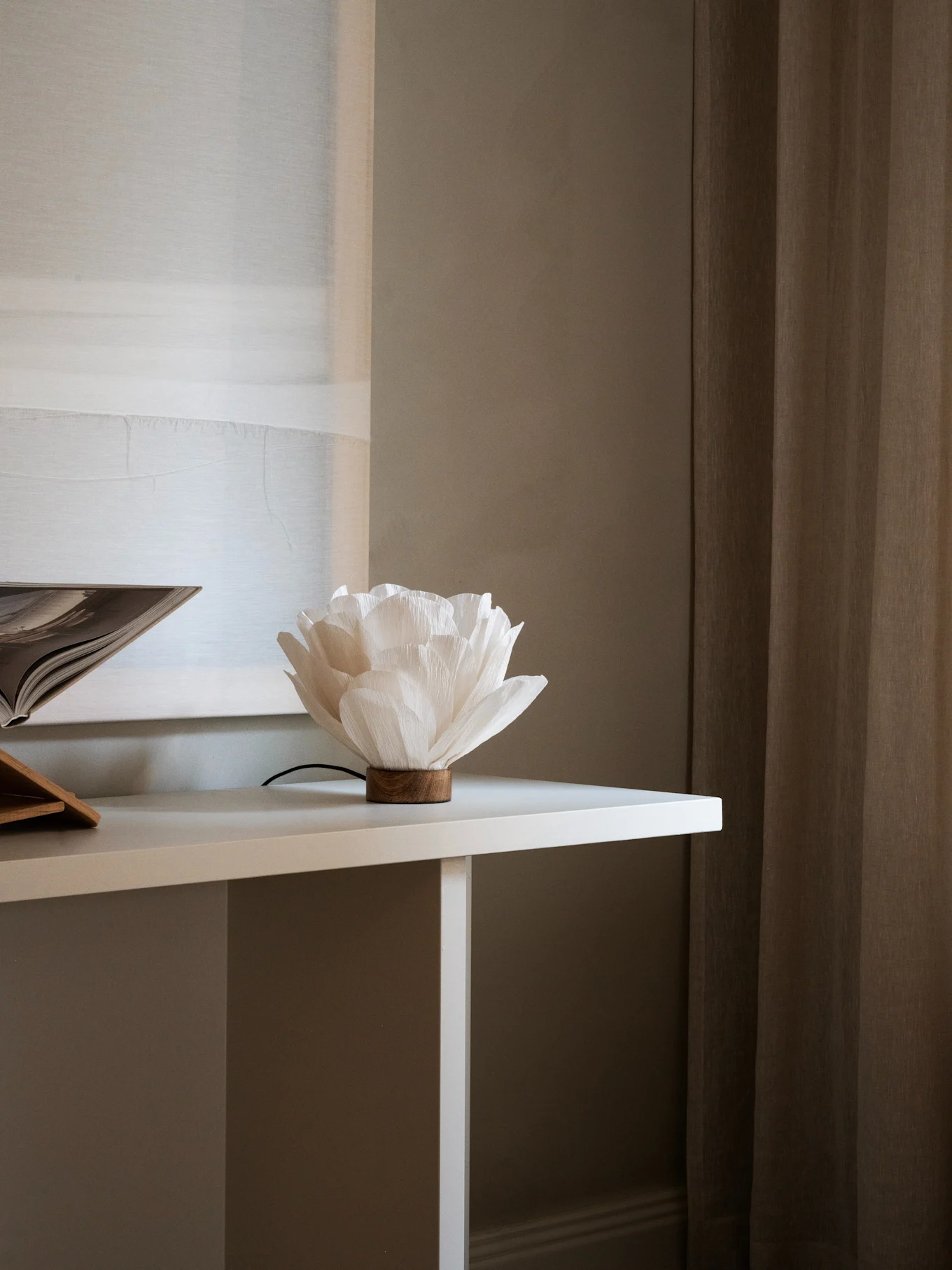 Lâmpada de papel branca em forma de flor com base de madeira numa mesa branca, ao lado de um livro aberto. Cortinas castanhas ao fundo.