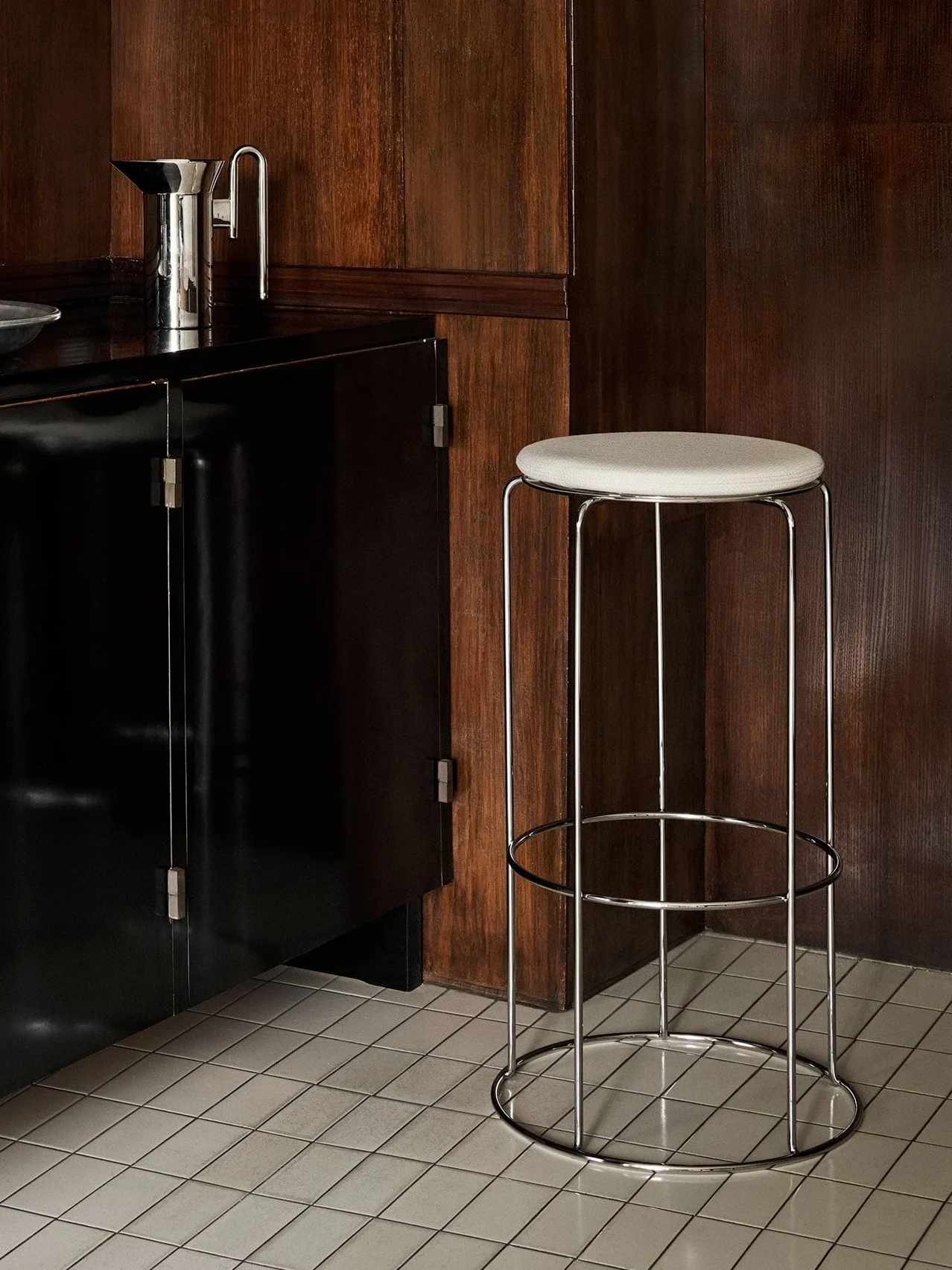 Um armário preto com utensílios de bar e um banco alto cromado com assento claro, contra painéis de madeira escura e chão de azulejos.
