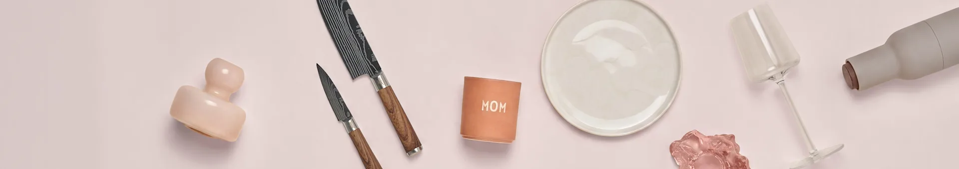 Cerâmica rosa, duas facas, chávena 'MOM', prato branco, vidro rosa, copo e garrafa cinzenta em superfície rosa.