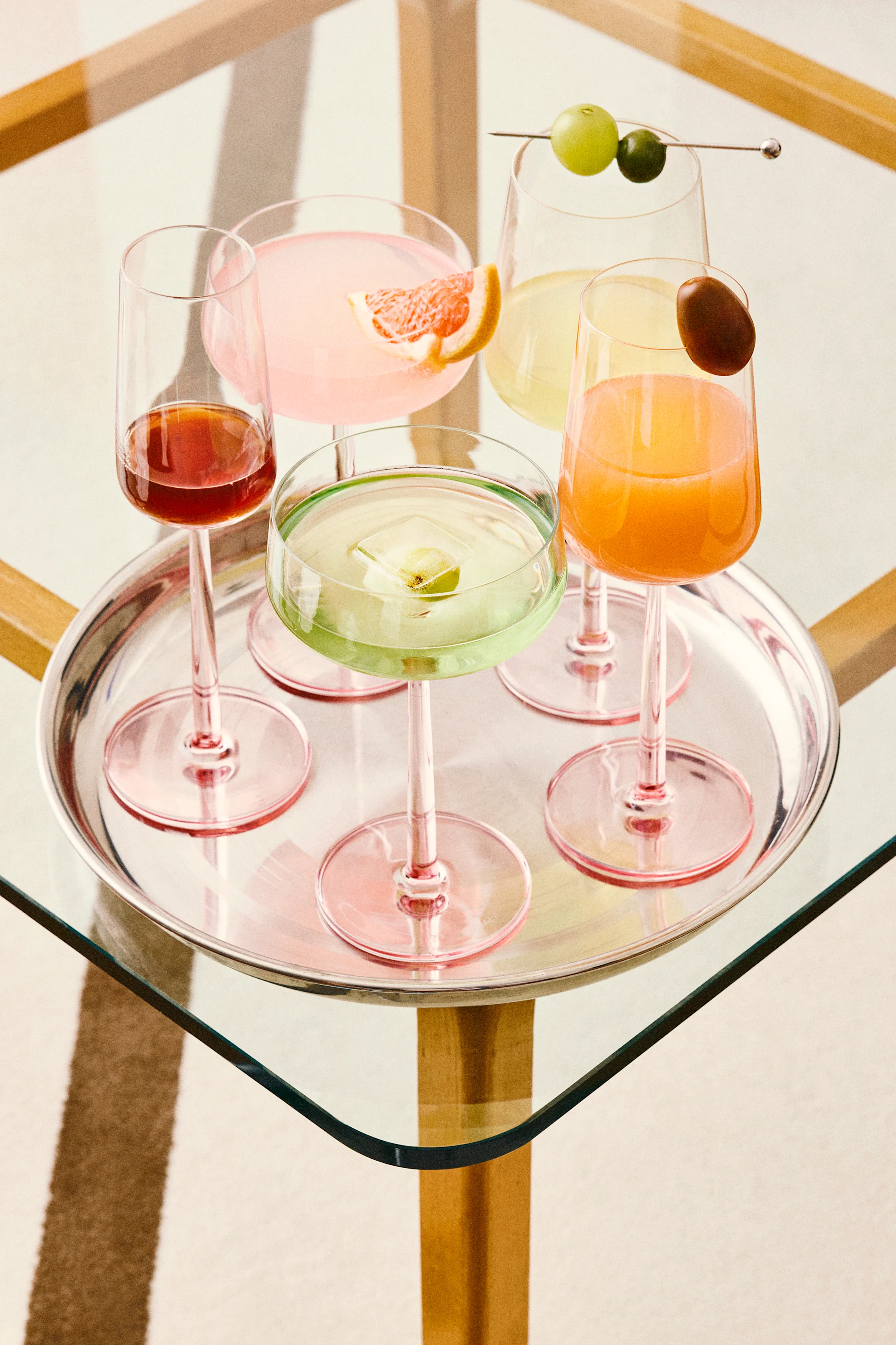 Cinco coquetéis coloridos em uma bandeja de prata, incluindo bebidas rosa, verdes, laranjas e vermelhas com várias guarnições, em uma mesa de vidro.