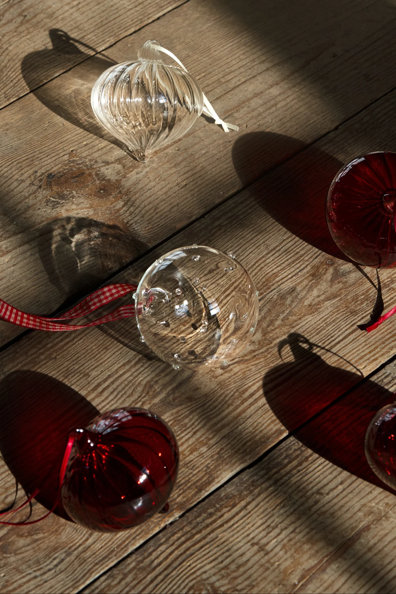Ornamentos de Natal de vidro transparentes e vermelhos com fitas espalhados sobre uma superfície de madeira ensolarada, criando sombras.