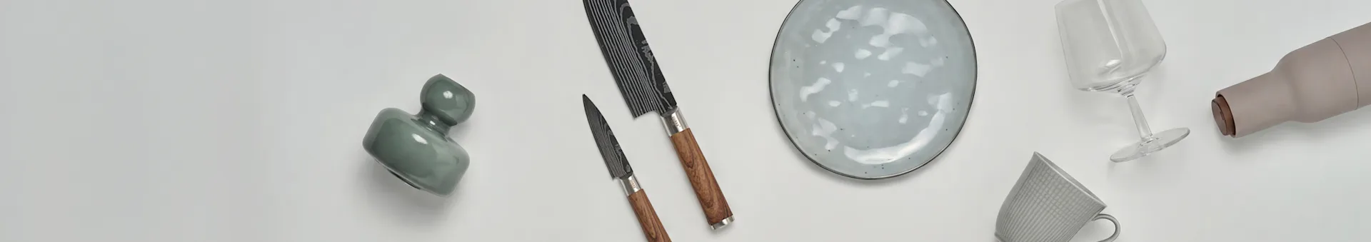 Vários itens de cozinha e jantar, incluindo um vaso verde, duas facas, um prato salpicado, uma taça de vinho, uma garrafa bege e uma caneca estampada.