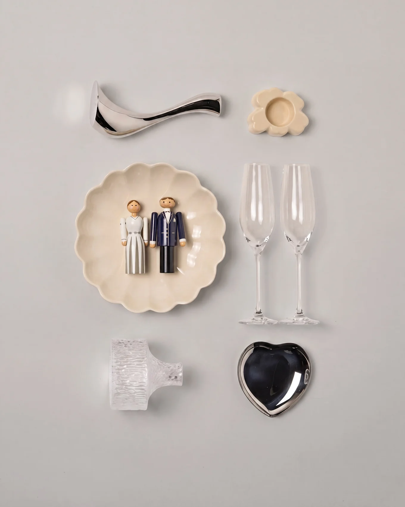 Disposição de casamento com figuras de madeira de noivos num prato em forma de concha, duas flutes de champanhe, um prato em forma de coração prateado e decorações variadas.