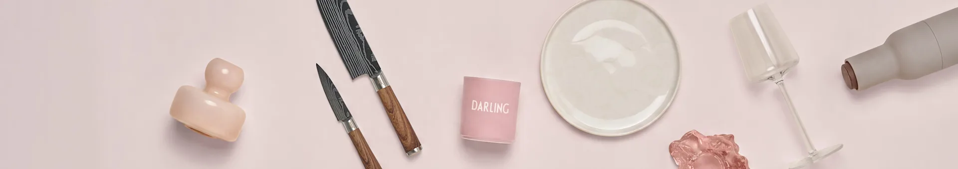 Sortido de artigos de cozinha e jantar em fundo rosa: uma escultura rosa, duas facas estampadas, um castiçal "DARLING", um prato creme, um copo de vinho transparente e uma garrafa cinzenta.