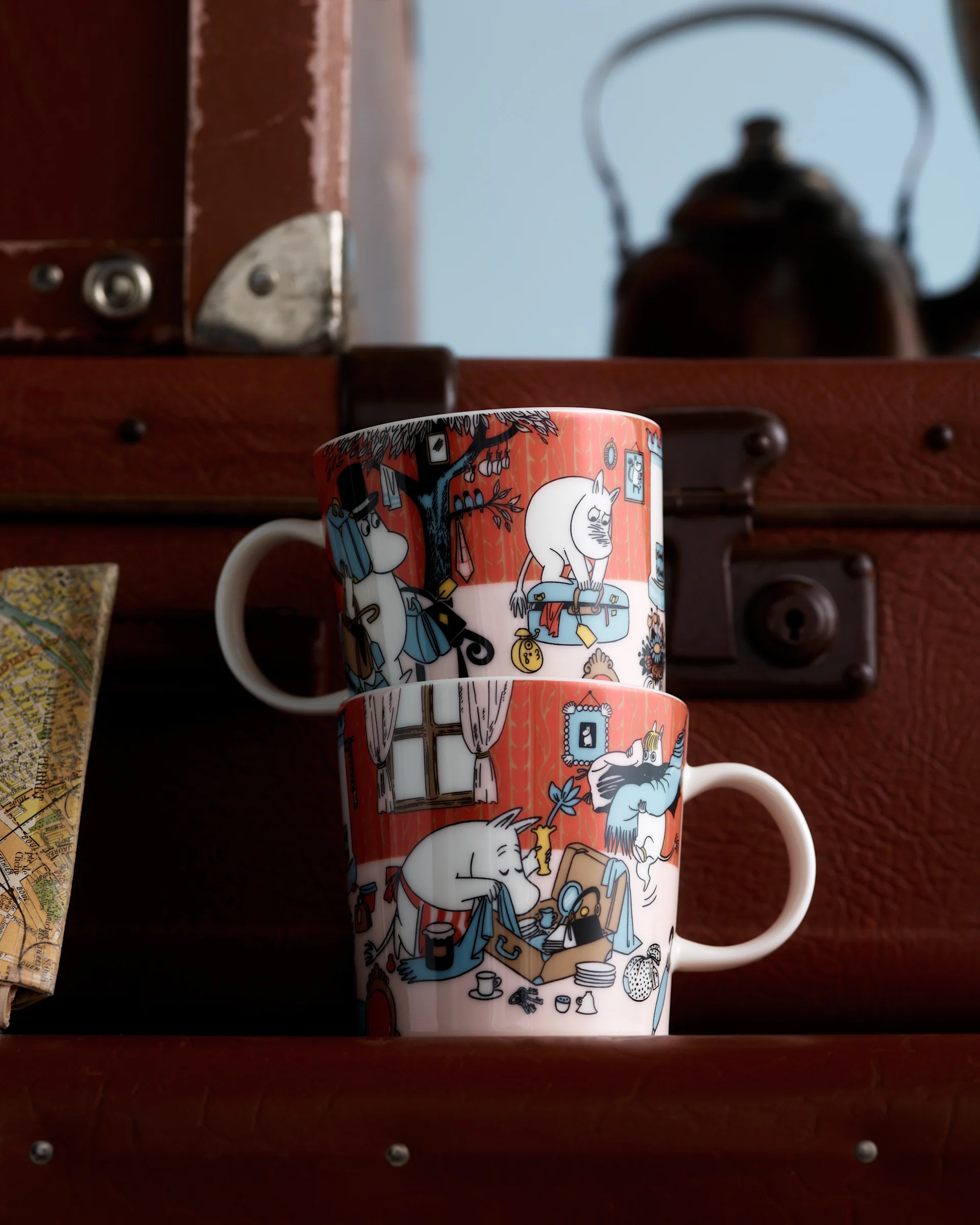 Duas canecas Moomin com ilustrações de personagens vermelho-alaranjadas, empilhadas à frente de malas castanhas e um mapa desfocado.